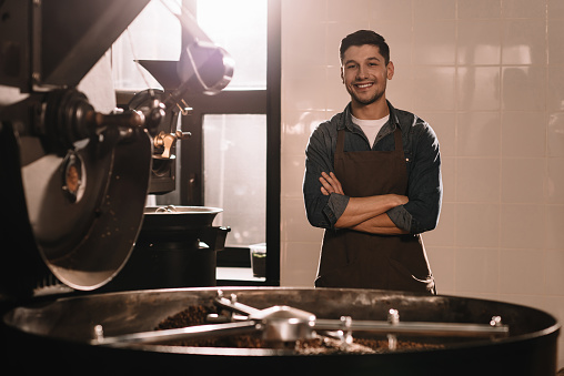 portrait of smiling coffee roaster standing at roasting machine and looking at camera