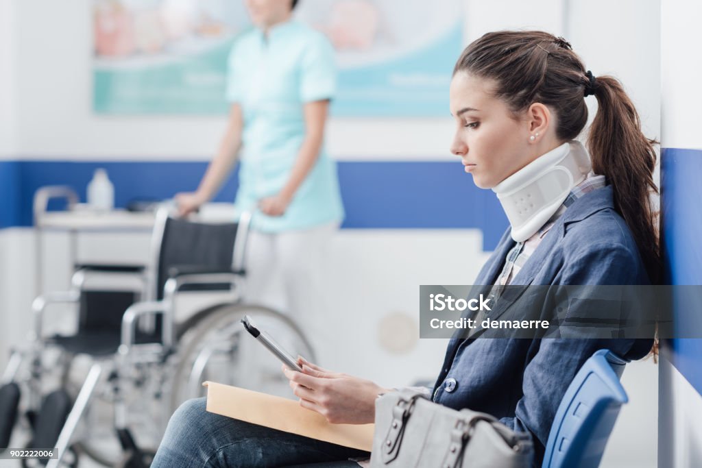 Young patient in the waiting room at the hospital Young female patient with cervical collar support at the hospital, she is sitting in the waiting room and connecting with a digital tablet, medical staff working on the background Physical Injury Stock Photo