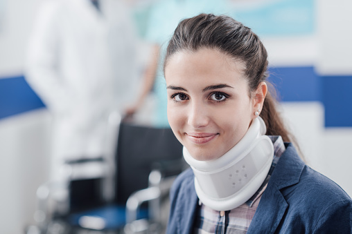 Young smiling patient at the hospital with cervical collar support and doctors working on the background