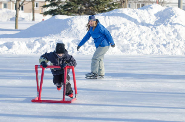 Young boy learning to ice skate using a helping accessory with mother behind