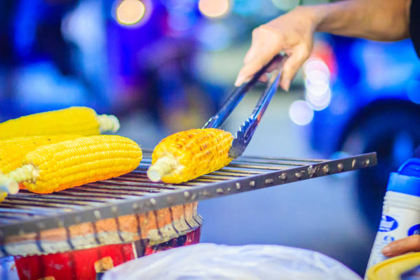 Close up hand of street food vendor while grilling for mixed sweet corn butter. Cook is grilling and sprinkling salt, sugar and butter to the grilled sweet corn on the electric stove.