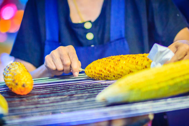 Close up hand of street food vendor while grilling for mixed sweet corn butter. Cook is grilling and sprinkling salt, sugar and butter to the grilled sweet corn on the electric stove.
