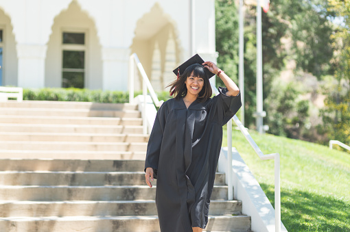 Attractive Filipino woman in graduation robe and cap walks down the  steps in celebration after graduation. She has a big smile and is walking toward the camera.