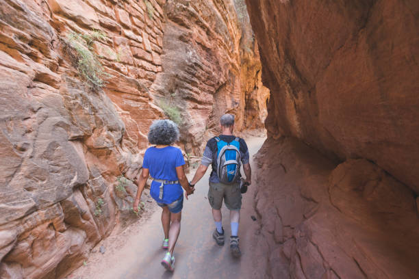 A couple in their 50s hikes through a majestic Utah canyon. They are surrounded on both sides by steep narrow, beautiful canyon walls. They are holding hands and the shot is from the rear.