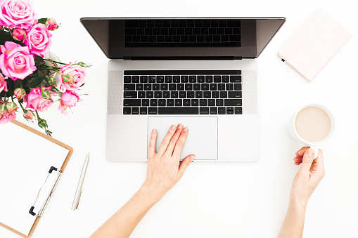 Woman freelancer working on laptop. Workspace with female hands, laptop, pink roses bouquet, coffee mug, diary on white table. Top view. Flat lay.