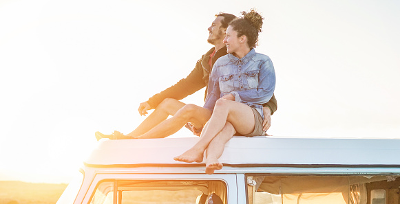 Happy couple sitting on top of minivan roof at sunset - Young people having fun on spring vacation traveling around the world - Travel,love and holiday concept - Focus on faces