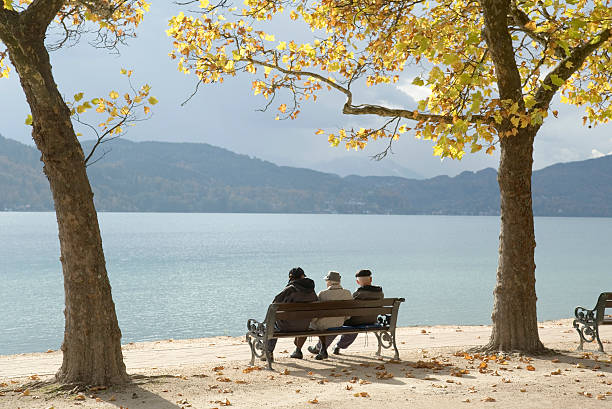 Three senior adults on lakeside bench under trees in autumn