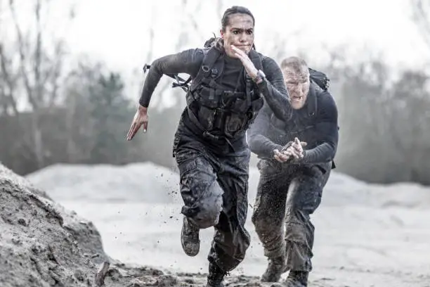 Photo of Redhead male drill instructor training beautiful brunette female soldier doing sprints outdoors in the mud