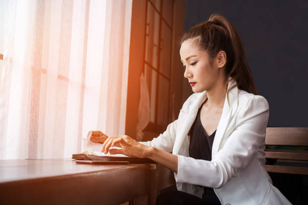 Asian beautiful in white with long brown hair businesswoman sitting reading a book account.worker receives an annual bonus.In coffee shop with sunset background. Asian beautiful in white with long brown hair businesswoman sitting reading a book account.worker receives an annual bonus.In coffee shop with sunset background. benefits of reading book stock pictures, royalty-free photos & images