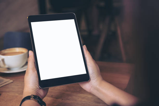 istock Mockup image of hands holding black tablet pc with blank white screen with coffee cup and mobile phone on wooden table in cafe 902564952