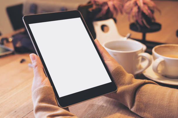 istock Mockup image of hands holding black tablet pc with blank white screen with coffee cups on wooden table in modern cafe 902565252