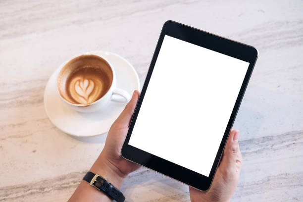 istock Top view mockup image of a woman's hands holding black tablet pc with blank white screen and white cup of latte coffee on marble table background 902568778