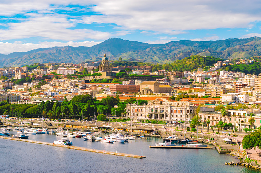 Cityscape of Messina, Sicily, Italy. Multi colored image with buildings, cathedral, mountains, blue sky with clouds, boats and yachts in the harbor or cruise port of Messina.
