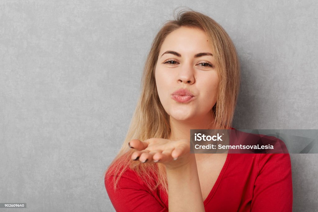 Horizontal shot of beautiful young woman with appealing appearance, makes air kiss, blows at camera, wears red sweater, isolated over grey bckground with copy space for your promotional text Adult Stock Photo