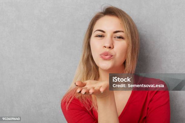 Horizontal Shot Of Beautiful Young Woman With Appealing Appearance Makes Air Kiss Blows At Camera Wears Red Sweater Isolated Over Grey Bckground With Copy Space For Your Promotional Text Stock Photo - Download Image Now