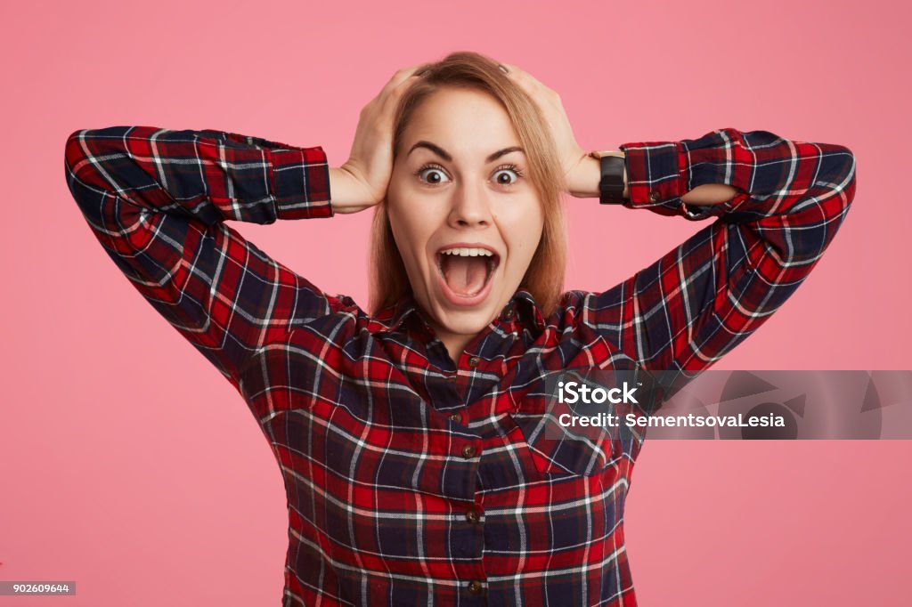 Frightened shocked young female opens mouth widely and exclaims in terror, doesn`t anticipate to see her phobia, keeps hands on head, poses against pink background in checkered stylish shirt Abundance Stock Photo