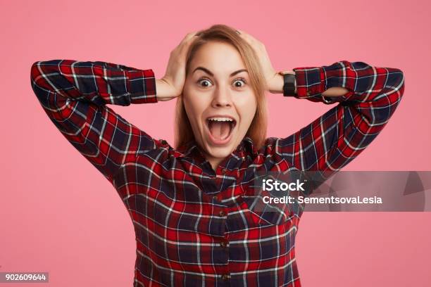 Frightened Shocked Young Female Opens Mouth Widely And Exclaims In Terror Doesnt Anticipate To See Her Phobia Keeps Hands On Head Poses Against Pink Background In Checkered Stylish Shirt Stock Photo - Download Image Now