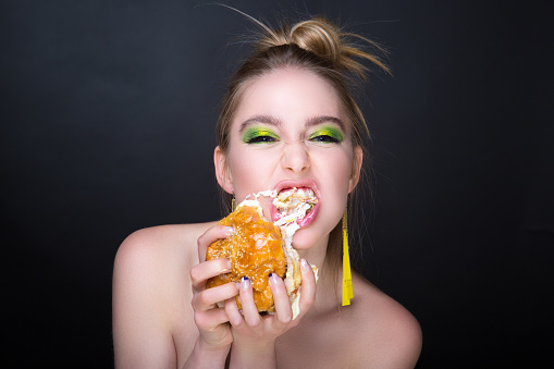 The woman is eating a giant hamburger with meat. Appetite lunch, delivery of food at home. Bright green fashionable make up eye shadows, yellow earrings. banner conceptual idea unhealthy food eating.