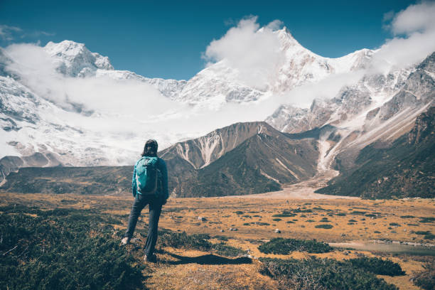 istock Standing woman with backpack looking on beautiful snow covered mountains in clouds at sunset. Landscape with girl, high rocks with snowy peaks, yellow grass, blue sky in Nepal. Travel. Vintage style 902683862
