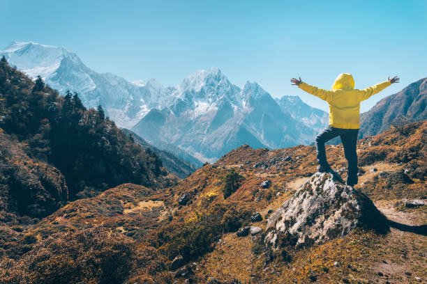 istock Standing man with raised up arms on the stone and looking on snow covered mountains. Landscape with traveler, high rocks with snowy peaks, grass, trees in autumn in Nepal. Lifestyle, travel. Trekking 902683910