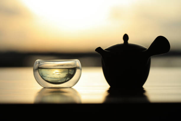 Glass teacup filled with Japanese green tea together with a kyusu teapot backlit on a polished wooden surface.
