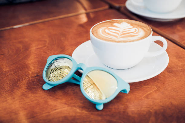 Close up white coffee cup  with heart shape latte art and sunglasses on wood table at cafe..