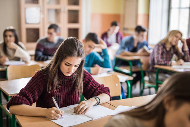 Female high school student writing a test in the classroom. Teenage girl taking notes while sitting in the classroom with her classmates. educational exam stock pictures, royalty-free photos & images