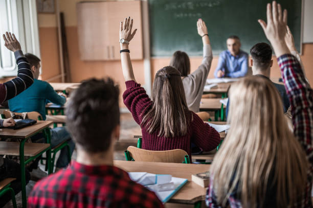 Rear view of high school students with raised arms during the class. Back view of group of students in the classroom raising hands to answer teacher's question. hand raised classroom student high school student stock pictures, royalty-free photos & images