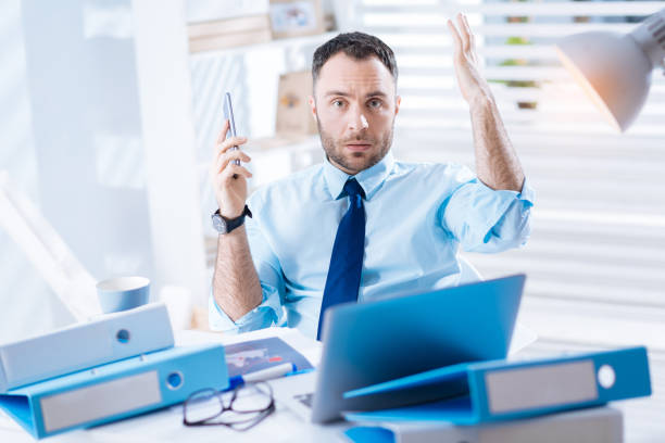istock Indignant young worker sitting with a smartphone and his hands up 902794822