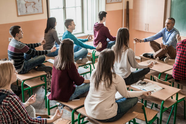 Group of students with their professor meditating in the classroom. Large group of high school students sitting in Lotus position with their teacher and practicing Yoga with their eyes closed. teen yoga stock pictures, royalty-free photos & images
