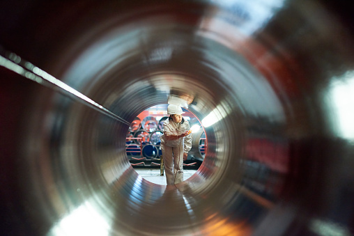 Busy female factory technician in hardhat focusing on work while examining tube at factory, view through object
