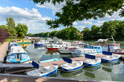 Joinville-le-Pont, Val-de-Marne, France - June 6, 2017: Riverboats, houseboats and electric rental boats mooring on the river Marne in the lovely marina of Joinville-le-Pont, in the inner suburb of Paris, by a sunny spring afternoon. The text on the small white and blue boats in the foreground says \
