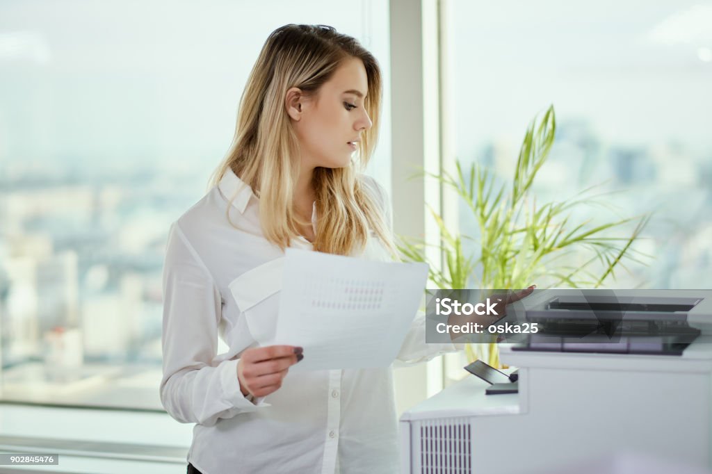 young businesswoman prints on the printer in the office beautiful young businesswoman poses in a sunny skyscraper office Printer - Occupation Stock Photo