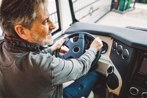 Mature man driving a camper, happy man having grey hair and a beard, wearing grey hoodie and dark blue trousers, holding a steering wheel, feeling happy, going on a trip, it's vacation time, road trips are the best.