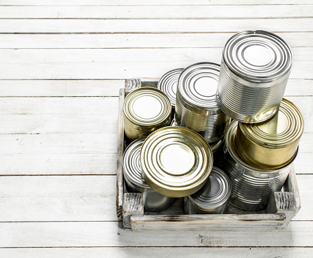 Tin cans with food in a box. On a white wooden table.