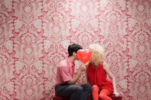 Top view of happy loving couple holding heart shape Valentines cards while leaning on bed surrounded with red balloons