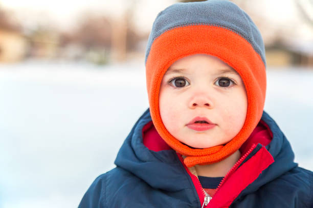 Close-up of a toddler (almost 18 months old) boy outside on a winter day. He is wearing a gray and orange fleece hat and winter jacket to keep him warm. He is looking at the camera with with big dark brown eyes.