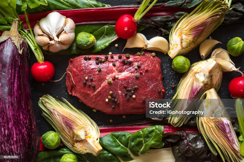 Delicious Home Cooking Piece of raw beef steak a slate table top background. The meat is seasoned with salt and pepper corns and surrounded by healthy vegetables such as Swiss chard (Ruby Red), garlic, Brussel sprouts, red radishes, artichoke and eggplant. Paleo Diet Stock Photo