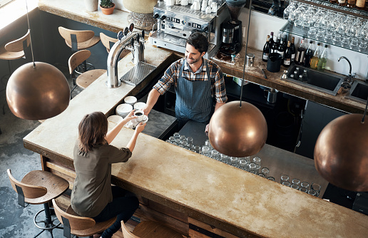 High angle shot of a cheerful young barman giving a coffee to a customer that she just payed for inside of a restaurant