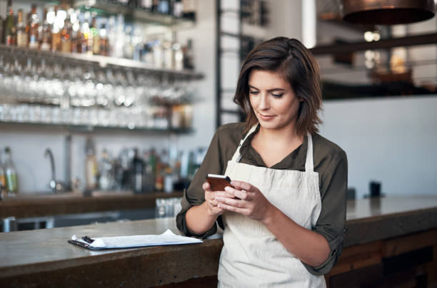 Making sure that everyone is coming to work today Shot of a cheerful young female bartender busy texting on her cellphone while standing next to the bar counter food service occupation photos stock pictures, royalty-free photos & images