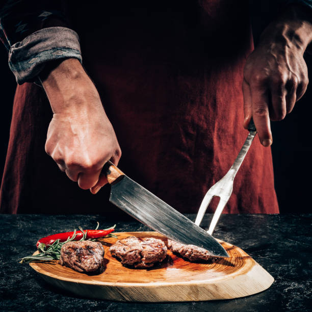 chef de close-up vue partielle dans le tablier avec la fourchette à viande et couteau tranche steak gourmet grillée au romarin et piment sur planche de bois - cooks knife photos et images de collection