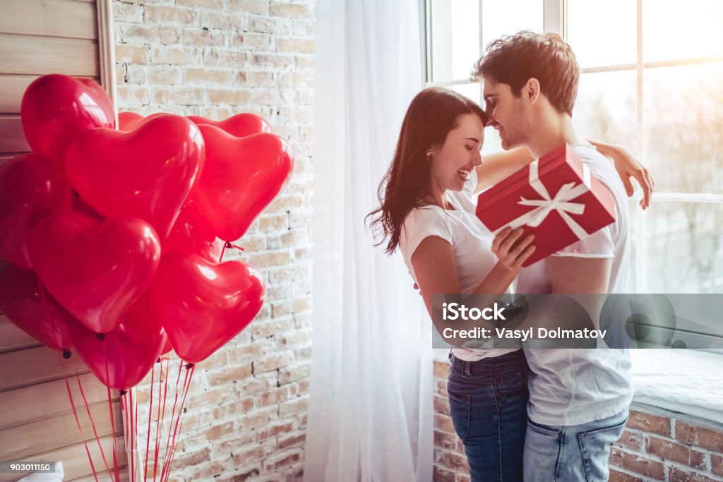 Couple in bedroom. Beautiful young couple at home. Hugging, kissing and enjoying spending time together while celebrating Saint Valentine's Day with gift box in hand and air balloons in shape of heart on the background. Valentine's Day - Holiday Stock Photo