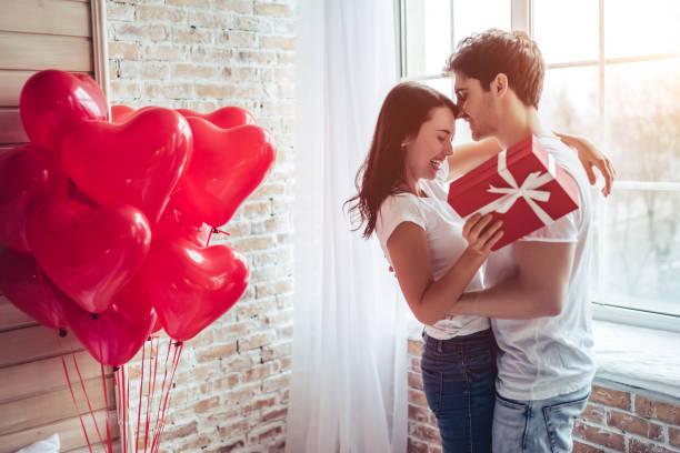Couple in bedroom. Beautiful young couple at home. Hugging, kissing and enjoying spending time together while celebrating Saint Valentine's Day with gift box in hand and air balloons in shape of heart on the background. valentines day holiday stock pictures, royalty-free photos & images