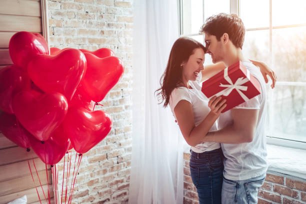 Couple in bedroom. Beautiful young couple at home. Hugging, kissing and enjoying spending time together while celebrating Saint Valentine's Day with gift box in hand and air balloons in shape of heart on the background. gift for girlfriend stock pictures, royalty-free photos & images