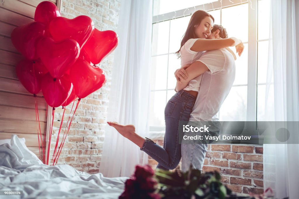 Couple in bedroom. Beautiful young couple at home. Hugging, kissing and enjoying spending time together while celebrating Saint Valentine's Day with red roses on bed and air balloons in shape of heart on the background. Valentine's Day - Holiday Stock Photo