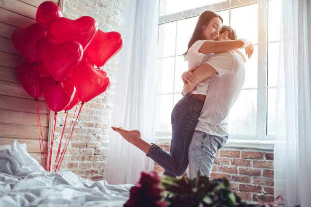 Couple in bedroom. Beautiful young couple at home. Hugging, kissing and enjoying spending time together while celebrating Saint Valentine's Day with red roses on bed and air balloons in shape of heart on the background. young man wink stock pictures, royalty-free photos & images