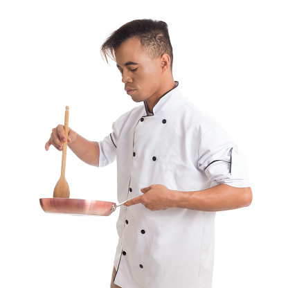 Professional is using a frying pan and wooden spoon. Young chef is in uniform. White background.