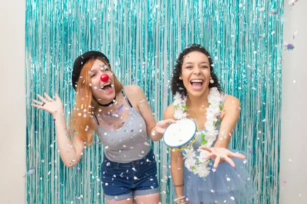 Two friends have fun at Carnaval. One is wearing clown nose and the other Hawaiian necklace. Predominance of color Blue. The friends laugh with the rain of confetti.