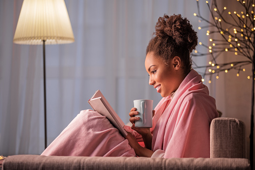 Side view of happy young woman reading book with interest. She is holding cup of coffee and smiling. Lady is sitting on sofa covered by blanket. Warm and comfort concept