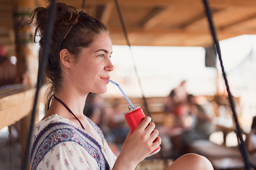 Attractive young woman relaxing on the swing at the beach bar and drinking soda.